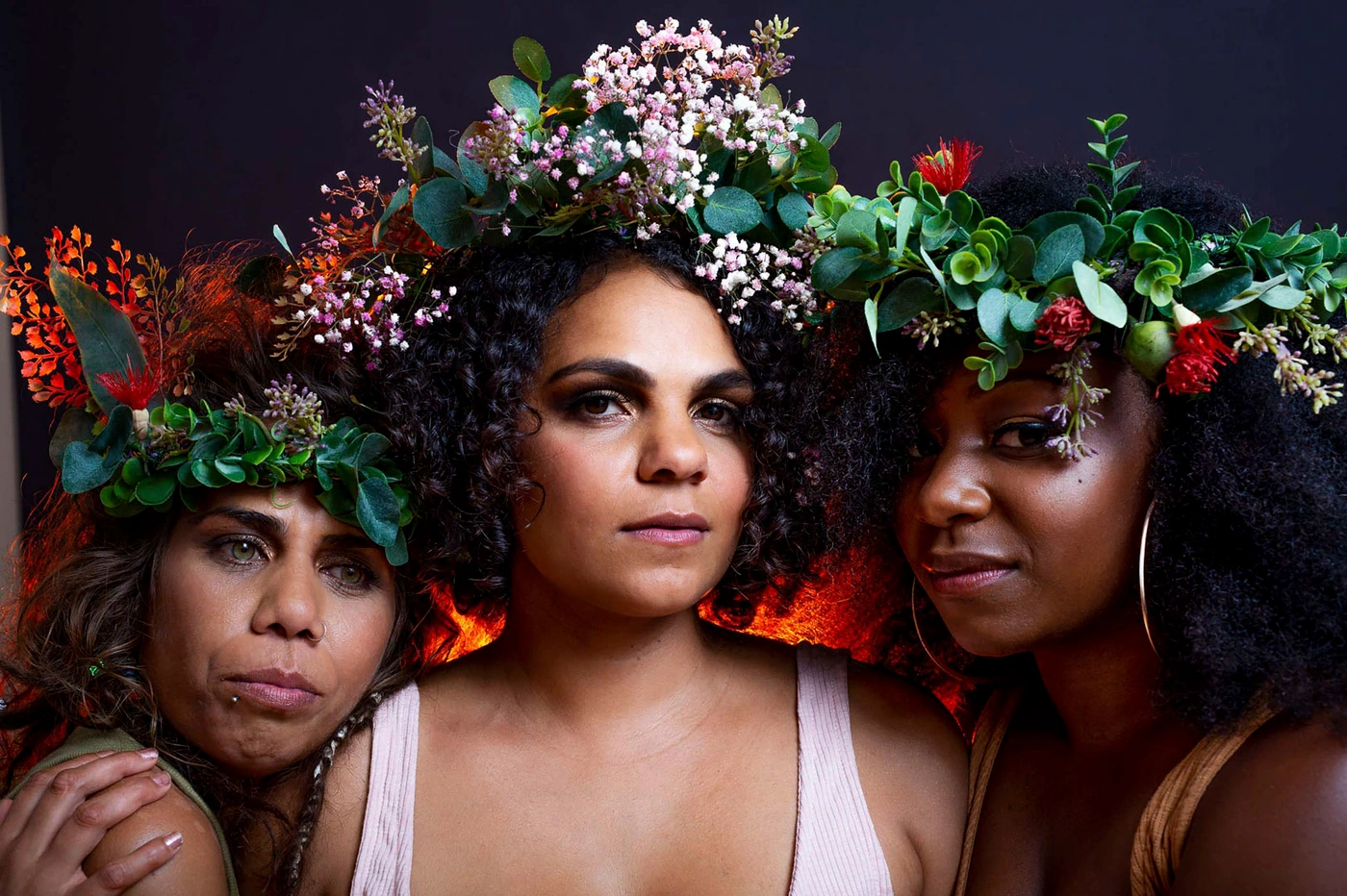 Three women wearing floral crowns and foliage headpieces posing together against a dark background in portrait photography by Mia Mala Mcdonald.