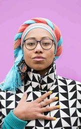 Portrait of a woman wearing a geometric patterned jacket, glasses, and a braided pink and turquoise headwrap against a pink backdrop