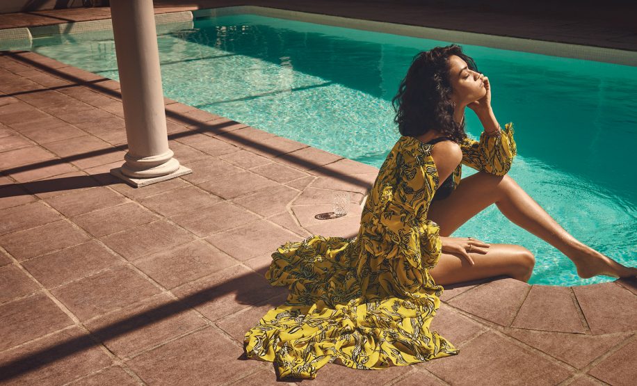 Woman in a yellow floral dress sits by a pool with columns, basking in the sun. Tiled patio and turquoise water create a relaxing scene.