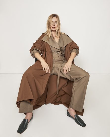 Fashion portrait of a blonde woman in brown coat and pants, seated in a studio with white background, wearing black loafers.