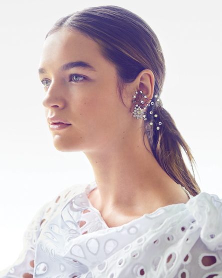 Close-up studio shot of a woman with light brown hair in a low ponytail, wearing a white lacy top and decorative earrings.