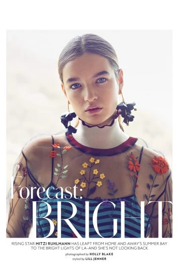 Portrait of Mitzi Ruhlmann wearing a sheer floral top and large black flower earrings, posing in front of a blurred window background.