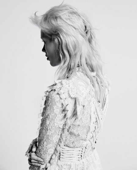 Profile portrait of a woman with light blonde, wavy hair wearing a white, lace dress, captured in black and white against a plain background.