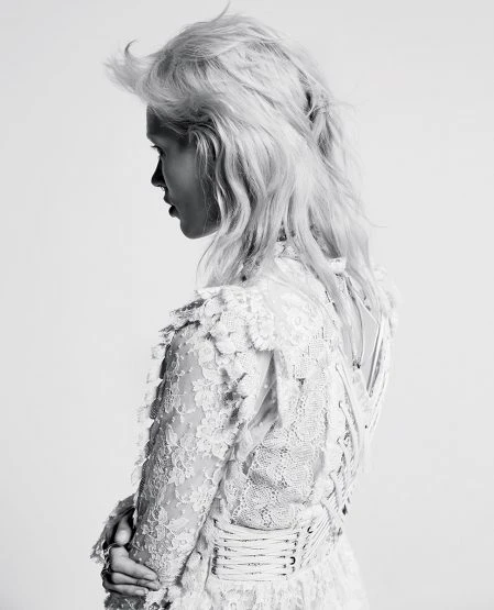 Profile portrait of a woman with light blonde, wavy hair wearing a white, lace dress, captured in black and white against a plain background.