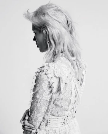 Profile portrait of a woman with light blonde, wavy hair wearing a white, lace dress, captured in black and white against a plain background.