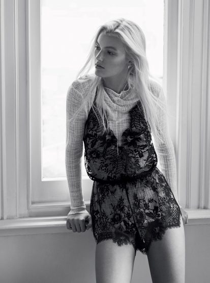 Black and white fashion shot of a blonde female model in a lace romper standing next to a window in an indoor setting.