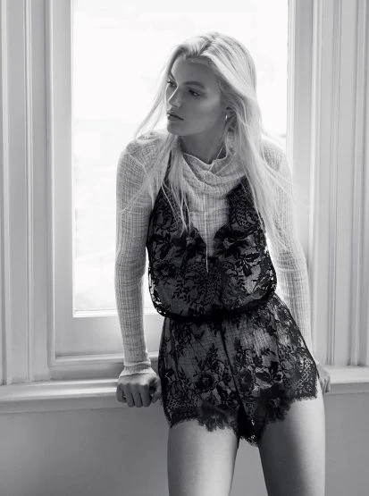 Black and white fashion shot of a blonde female model in a lace romper standing next to a window in an indoor setting. 