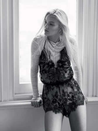 Black and white fashion shot of a blonde female model in a lace romper standing next to a window in an indoor setting. 