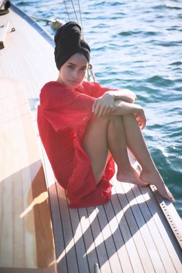 A young woman with a black head covering sits on the wooden deck of a yacht in a red dress with the ocean in the background.