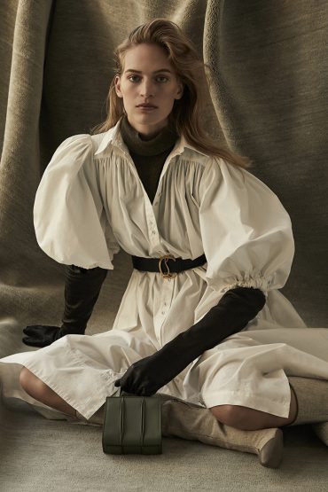 Elegant woman poses cross-legged indoors wearing a white dress with puffed sleeves, black gloves, and a structured olive-green handbag.