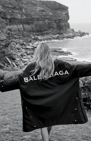 Woman wearing a Balenciaga jacket outdoors with an ocean and rocky cliff background in a black and white fashion photograph.