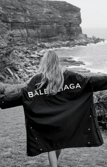 Woman wearing a Balenciaga jacket outdoors with an ocean and rocky cliff background in a black and white fashion photograph.