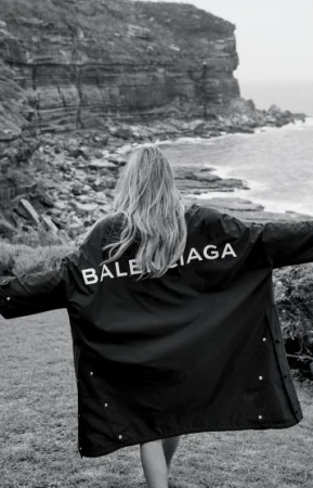 Woman wearing a Balenciaga jacket outdoors with an ocean and rocky cliff background in a black and white fashion photograph.