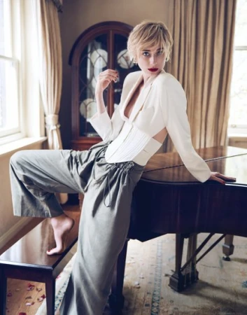 Short-haired blonde woman posing by a grand piano wearing a white structured blazer and grey trousers.