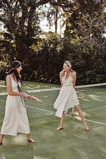 Two women in white outfits playing tennis on a green court surrounded by trees, enjoying an outdoor daytime activity.