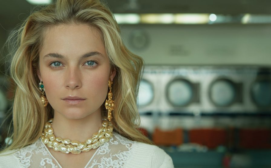 Close-up of a blonde adult female wearing a pearl necklace and gold earrings in front of commercial washing machines.