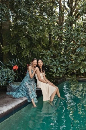 Two women sitting on the edge of an outdoor pool with their feet in the water, surrounded by lush green tropical foliage.