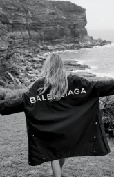 Black and white image of a woman from behind wearing a branded coat with outstretched arms, outdoors near rocky cliffs and water.