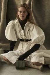 Model seated wearing a white puffed-sleeve dress, dark gloves, and holding a small green handbag against a textured fabric backdrop