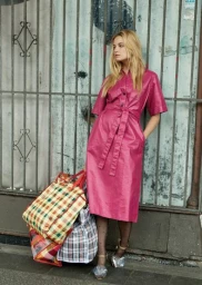 Blonde woman in magenta dress leaning against weathered iron grating with plaid market bags