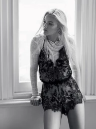 Monochrome portrait of a blonde woman wearing a sheer patterned top and dark lace shorts standing by a window