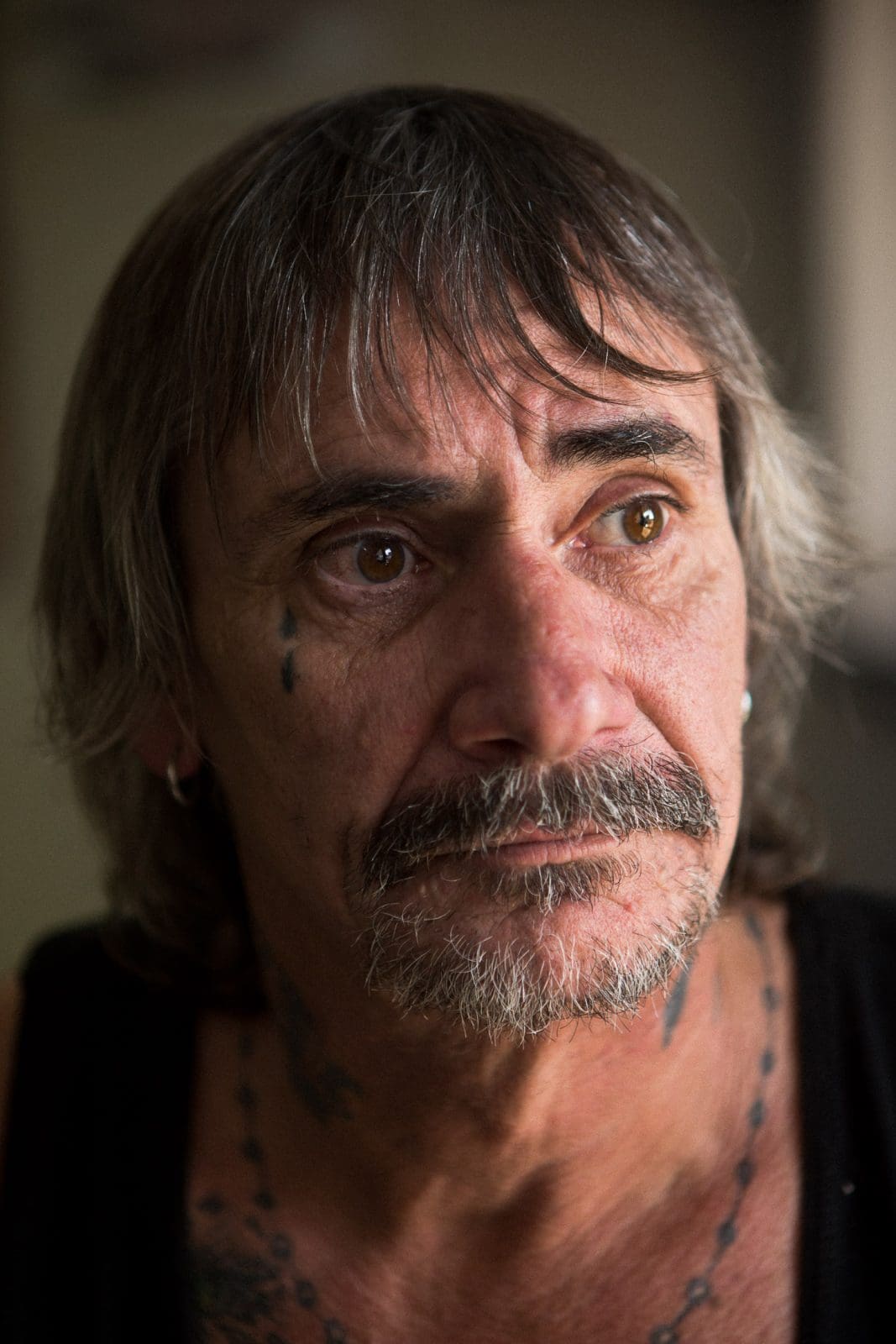 Close-up portrait of a mature man with brown hair, a mustache, and tattoos, captured indoors with natural lighting and shallow depth of field.