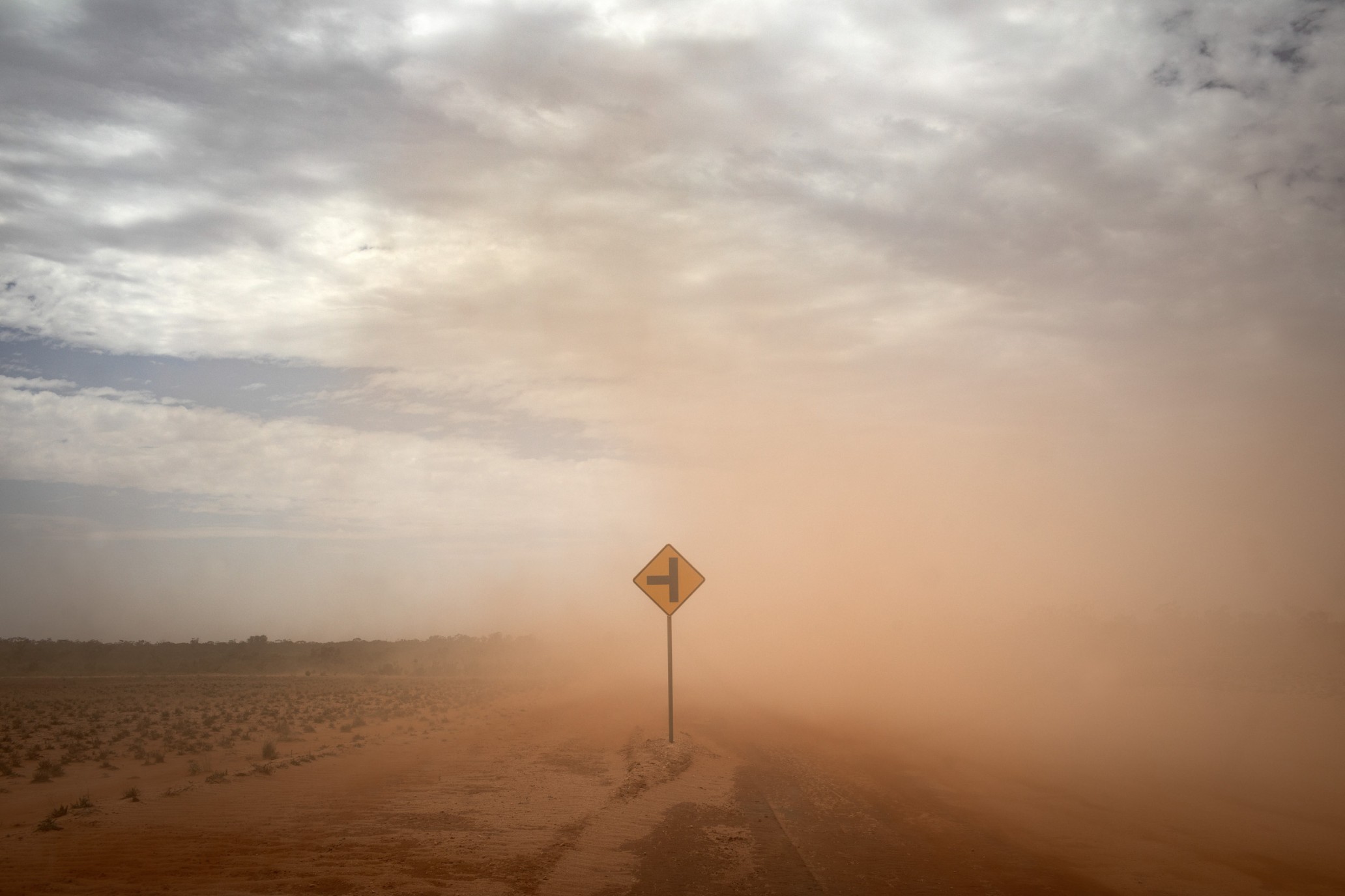 A rural road scene with a yellow traffic sign, enveloped in a thick, reddish-brown dust storm, under an overcast sky with light patches.