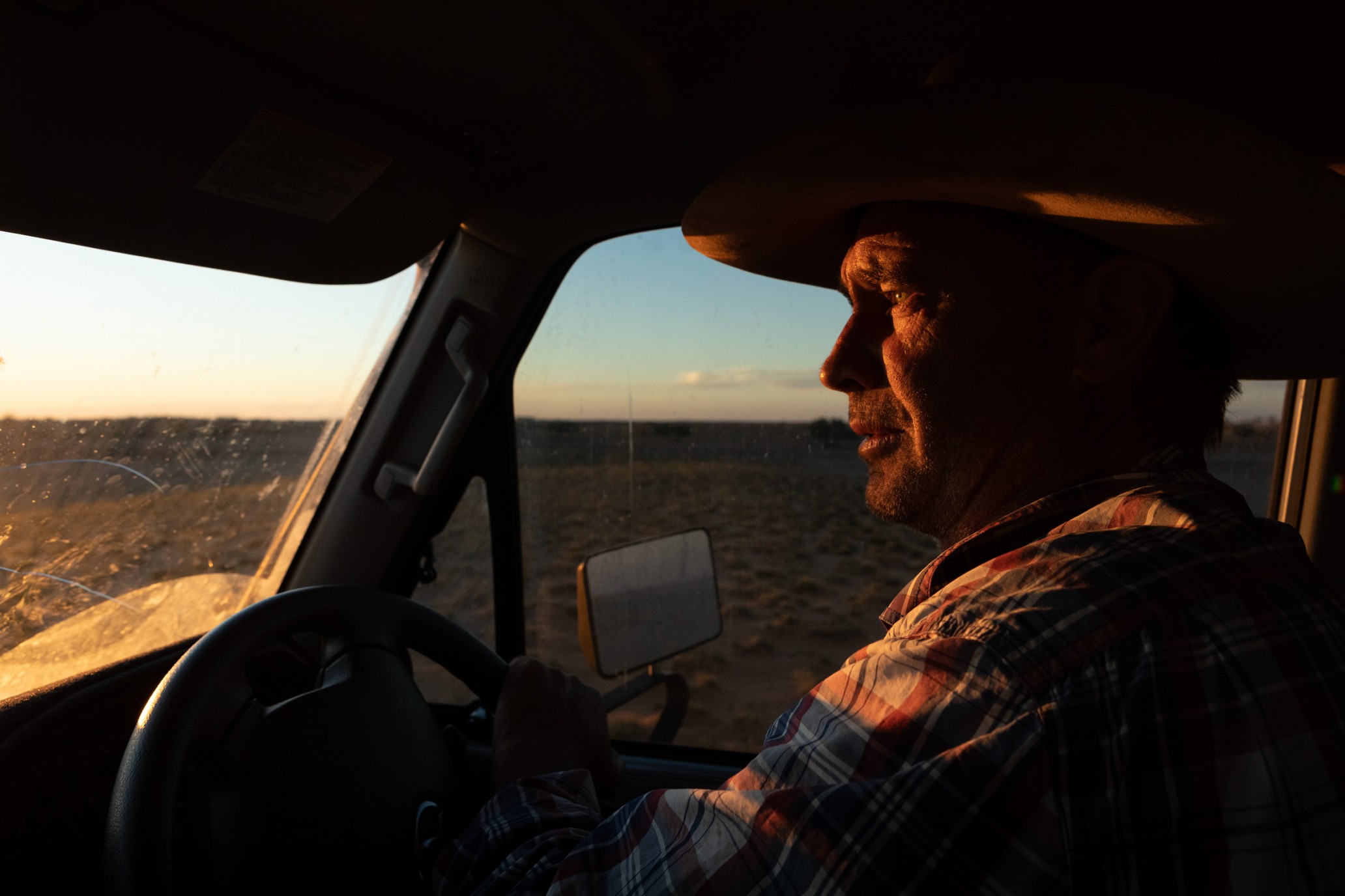 Man in a cowboy hat driving a car at sunset in a desert landscape. Golden light shines on his face as he holds the steering wheel.