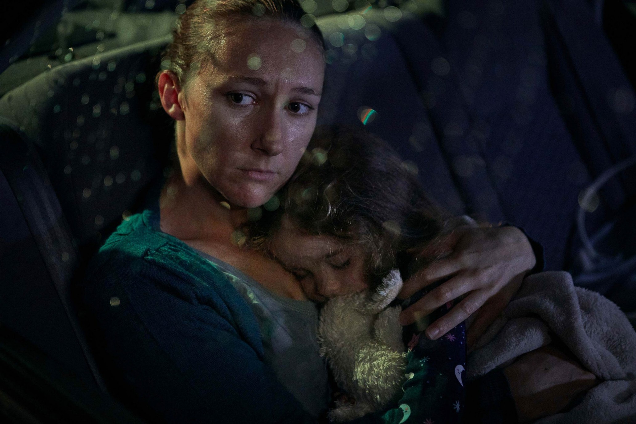 A mother and her young child, embracing tightly, are seated in the dim interior of a car, with bokeh lights visible in the background.