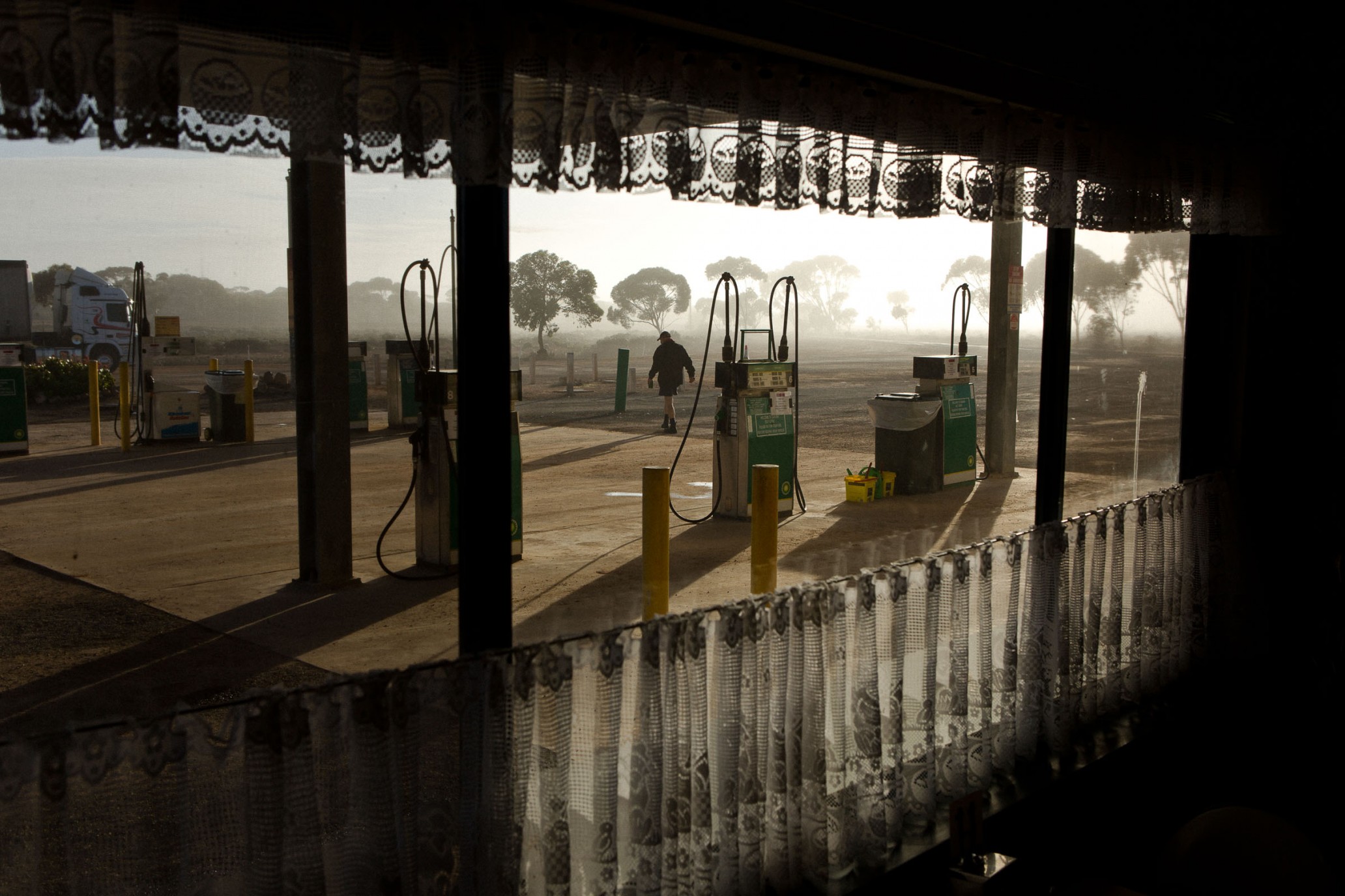 A rural gas station scene with vintage pumps, observed through an ornate curtained window, bathed in the glow of early morning sunlight.