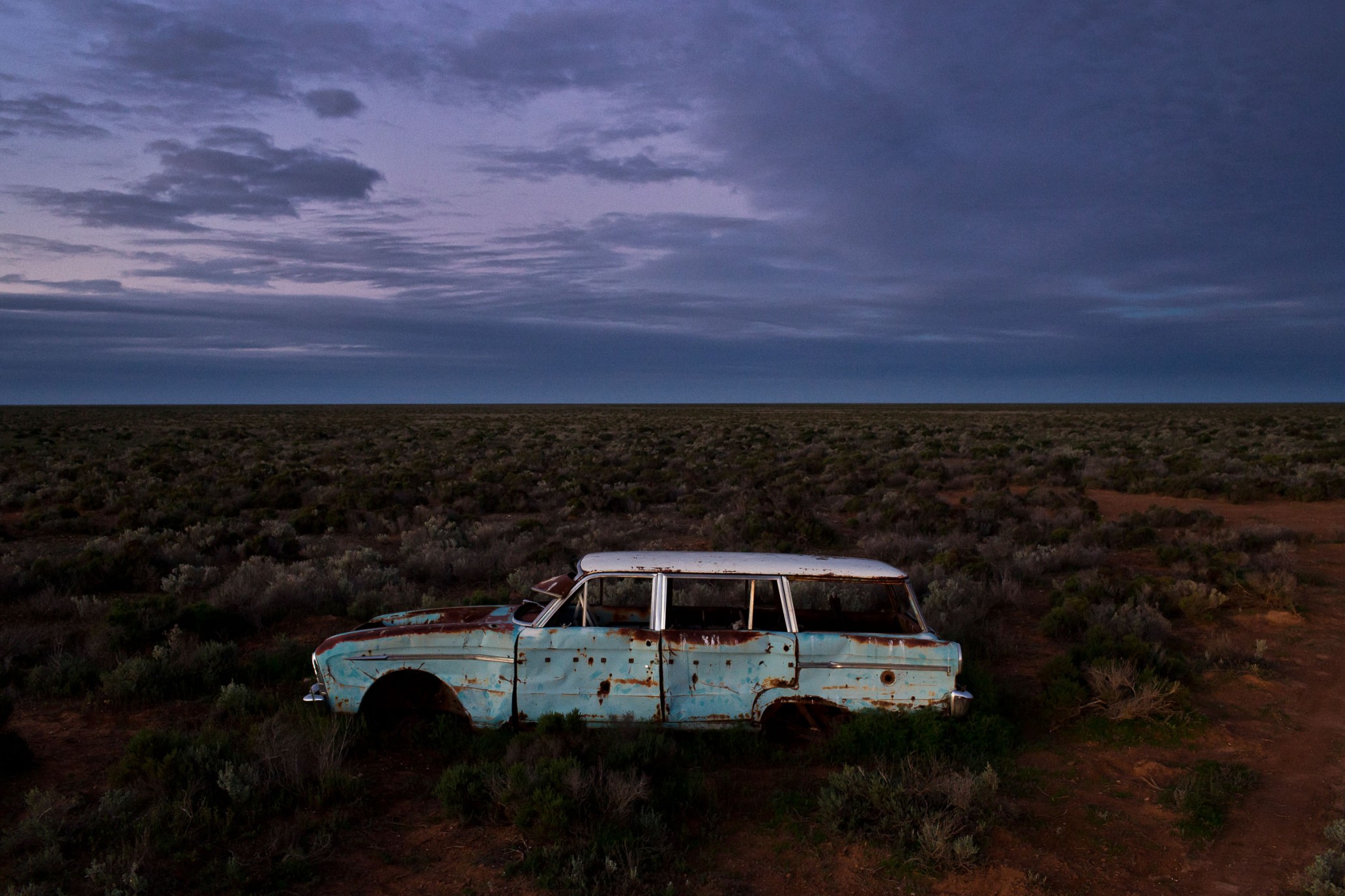 An abandoned, rusted car sits in a desolate field under a cloudy sky, evoking a sense of decay and forgotten history in the landscape.