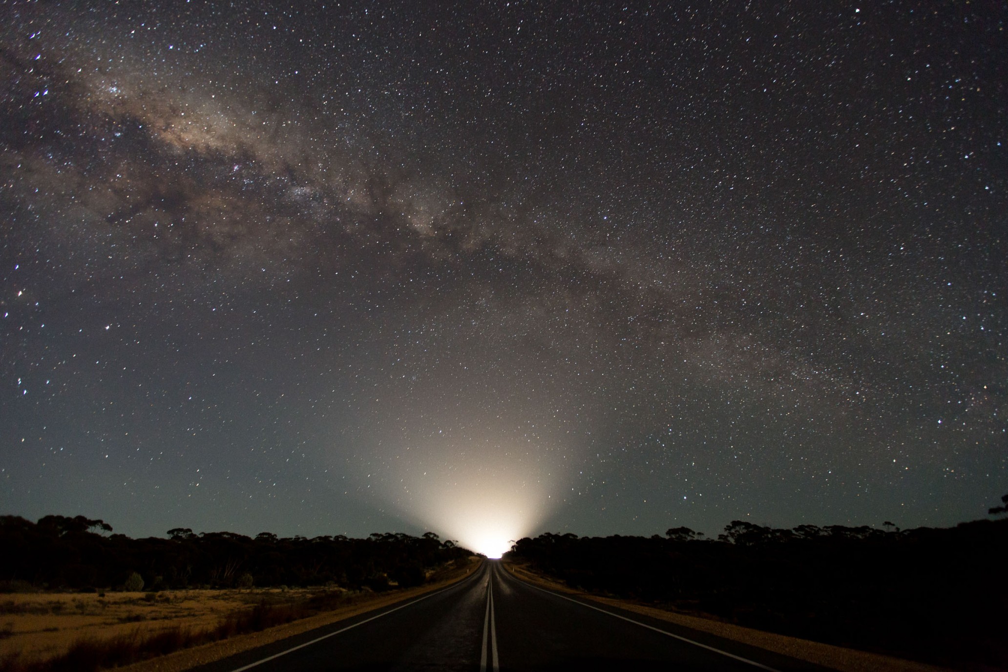 A scenic nightscape features a long road illuminated by distant lights under a clear sky filled with stars, including the Milky Way.