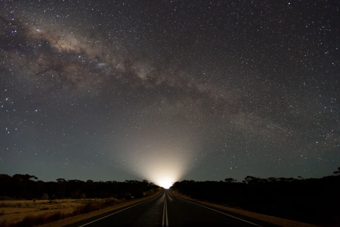 A scenic nightscape features a long road illuminated by distant lights under a clear sky filled with stars, including the Milky Way.
