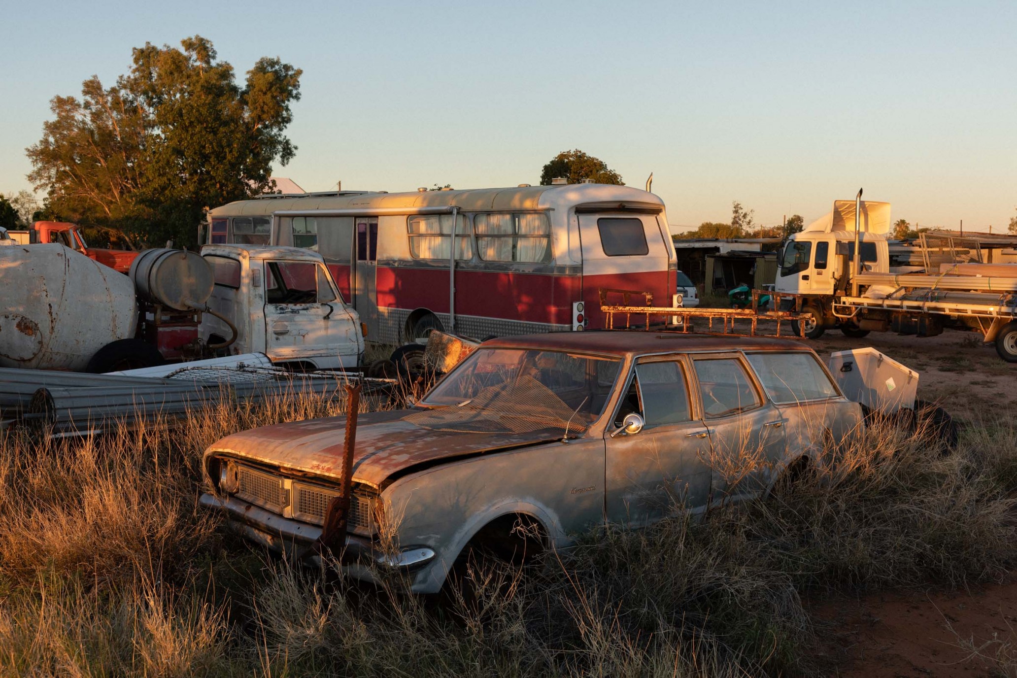 Vintage station wagon car in disrepair, surrounded by tall grass, with a bus and truck in the background under a clear sky at dusk.