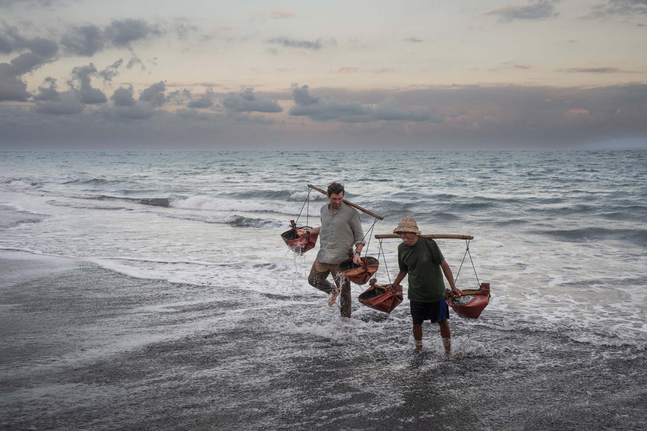 Two men wade through surf on a beach carrying buckets balanced on shoulder poles under an overcast sky at dusk.