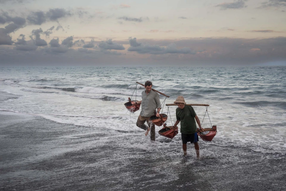 Two men wade through surf on a beach carrying buckets balanced on shoulder poles under an overcast sky at dusk.