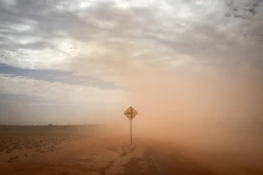 A dust storm partially obscures a road sign and a flat, dry landscape under an overcast sky.
