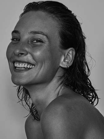 Close-up portrait of a smiling woman with slicked-back wet hair and hoop earrings against a neutral grey background