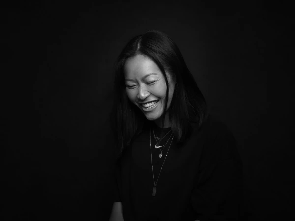 Portrait of a laughing east asian woman wearing layered necklaces against a solid black background by Jez Smith.