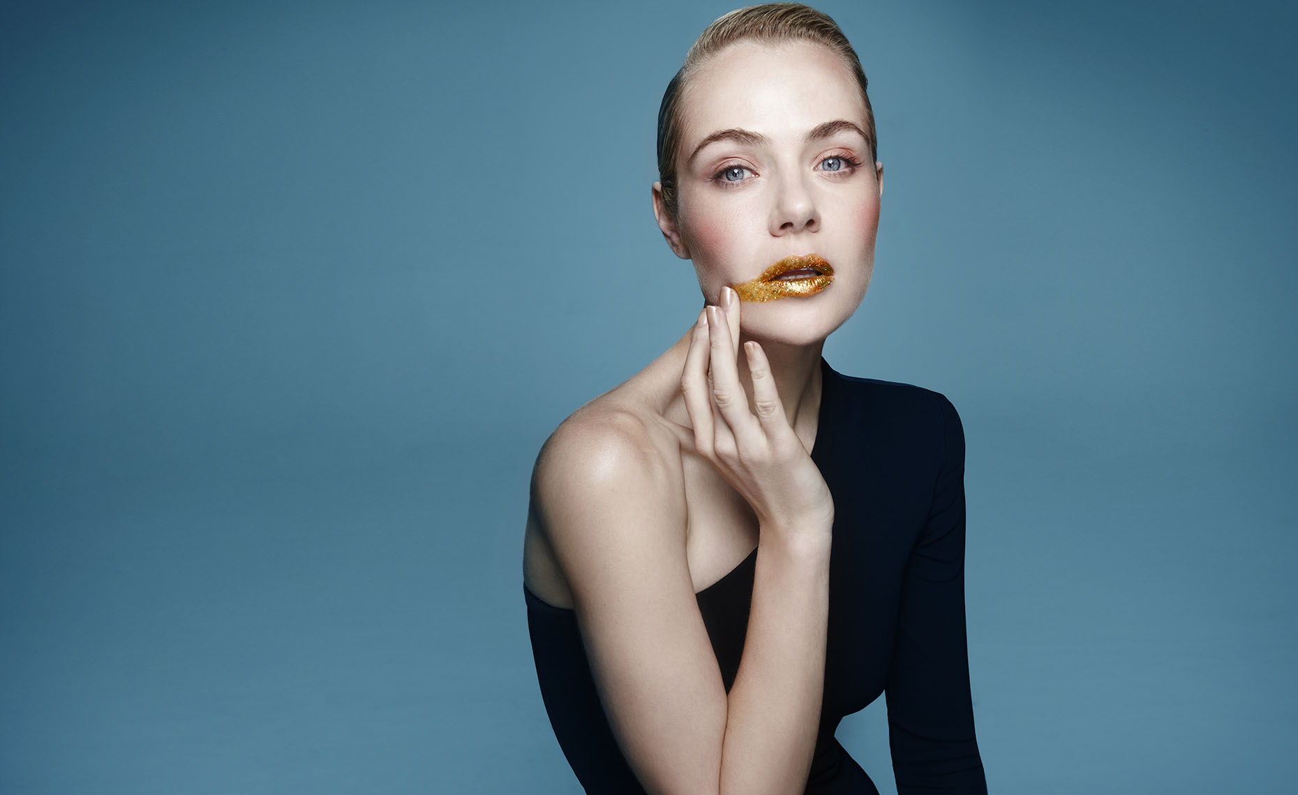 Elegant studio portrait of a blonde woman with gold glitter lips, wearing a black dress, posing against a blue background.
