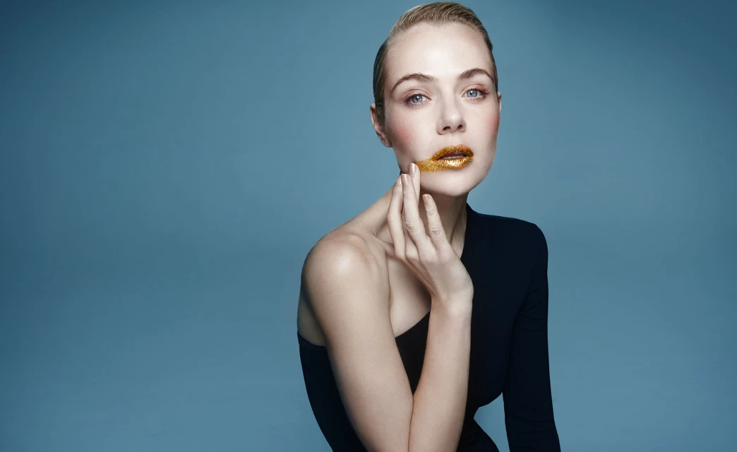 Elegant studio portrait of a blonde woman with gold glitter lips, wearing a black dress, posing against a blue background.
