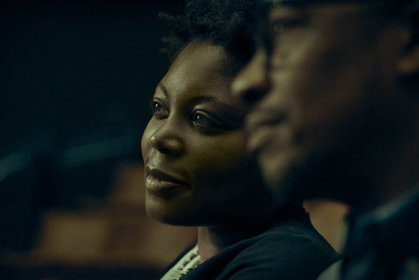 Close-up of an adult Black woman and man in a dimly lit setting, both looking to the side with soft lighting highlighting their faces.
