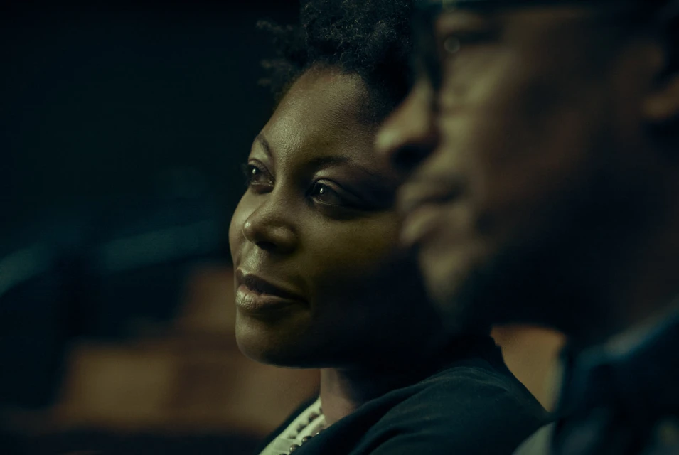 Close-up of an adult Black woman and man in a dimly lit setting, both looking to the side with soft lighting highlighting their faces.