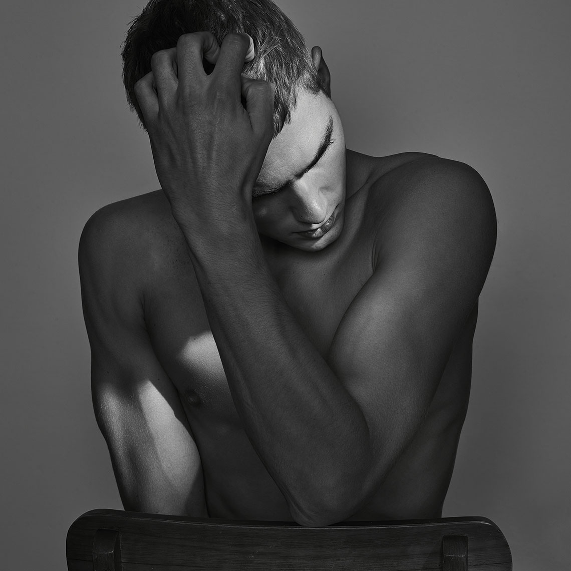 Black and white portrait of a shirtless adult male sitting on a chair with his hand on his head, looking down in a photo studio.