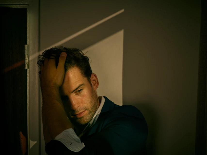 Adult male in a dark suit poses in a shadowed corner with his hand on his head in a dimly lit room.