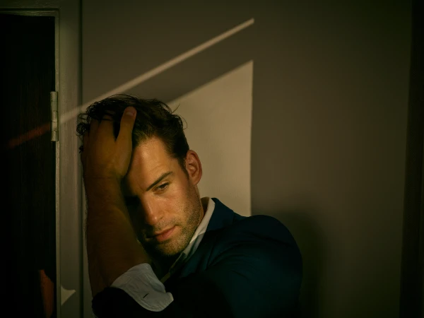 Adult male in a dark suit poses in a shadowed corner with his hand on his head in a dimly lit room.