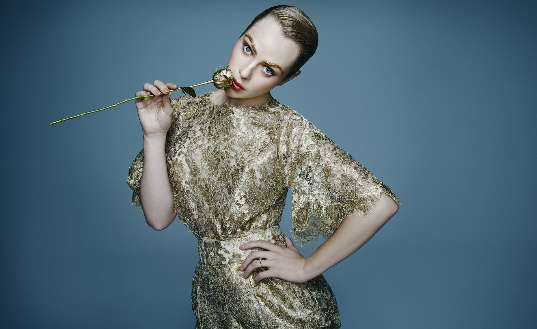 Blonde woman in a gold lace dress holds a gold rose against a blue backdrop, striking a pose with hand on hip.