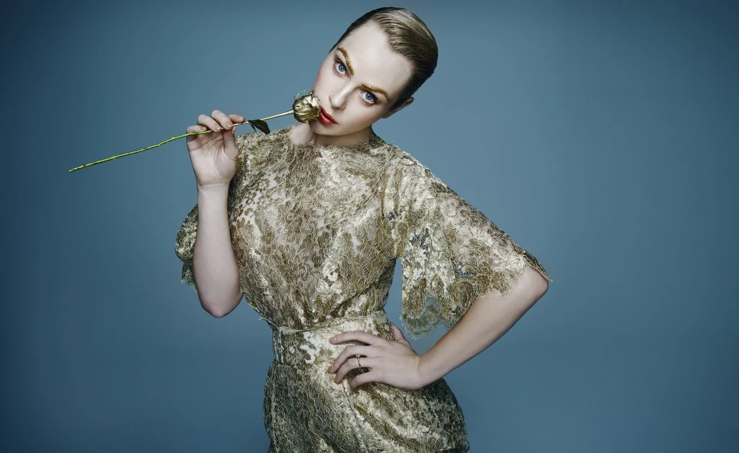 Blonde woman in a gold lace dress holds a gold rose against a blue backdrop, striking a pose with hand on hip.