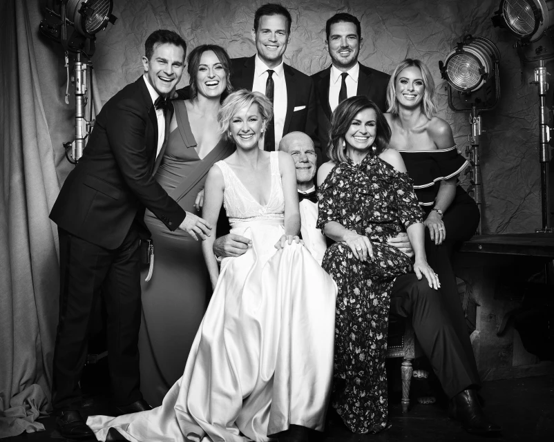 Black and white photo of a group of eight adults posing in formal attire in a photo studio with vintage spotlights in the background.