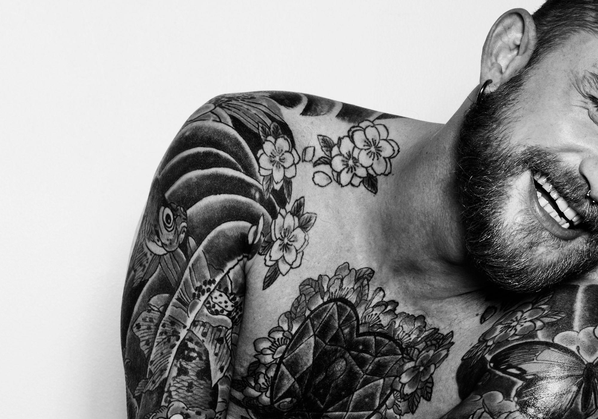 Black and white portrait of a smiling man with a beard and extensive tattoos covering his upper body against a white background.